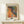 May include: Framed art print featuring vibrant abstract design with colorful polka dots and organic patterns in orange, yellow, blue, and earth tones, displayed on light wood shelf beside small wrapped gift with twine and greenery sprig against cream wall.