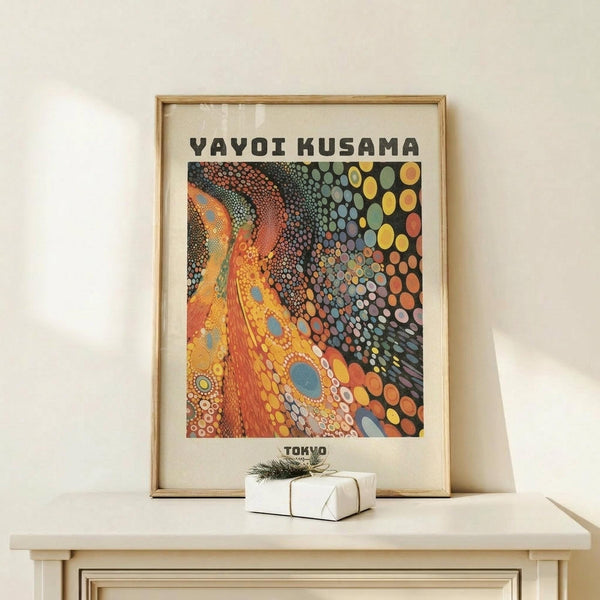 May include: Framed art print featuring vibrant abstract design with colorful polka dots and organic patterns in orange, yellow, blue, and earth tones, displayed on light wood shelf beside small wrapped gift with twine and greenery sprig against cream wall.