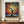 May include: Framed art print featuring a dark silhouetted tree with sprawling branches against a vibrant cosmic background of multicolored dots and circles in warm oranges, blues, and yellows. The artwork has Japanese text below the tree image. The brown wooden frame sits on a light wood credenza next to a white ceramic vase and stack of books, against cream walls with white molding.