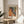 May include: Framed art print featuring Yayoi Kusama's colorful dot artwork in warm oranges, yellows, and cool blues with "TOKYO" text below, displayed on a light wood round side table next to a cream upholstered chair, with plant shadows cast on white walls from natural window light.