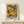 May include: Framed poster print featuring colorful abstract floral patterns in orange, green, yellow, and white dots and circles against dark background, displayed on light wood shelf with small wrapped gift box beside it on cream wall