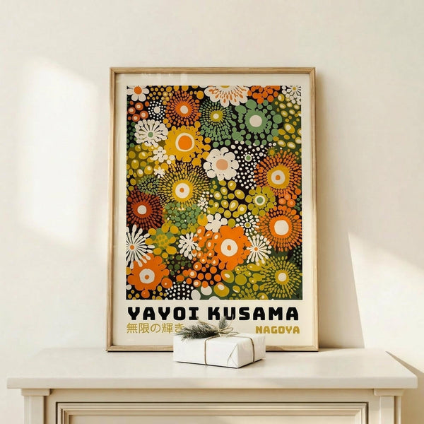May include: Framed poster print featuring colorful abstract floral patterns in orange, green, yellow, and white dots and circles against dark background, displayed on light wood shelf with small wrapped gift box beside it on cream wall