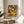 May include: Framed poster featuring vibrant circular floral patterns in orange, yellow, green, and white dots displayed on a light wood round side table next to a beige upholstered chair. Natural sunlight streams across neutral walls with plant leaves visible in foreground. The artwork shows densely packed polka-dotted flowers and geometric designs with Japanese text at bottom in thin wooden frame.