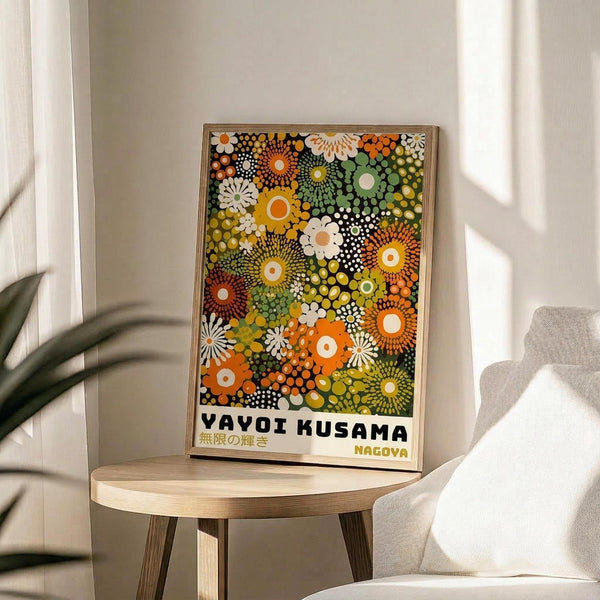 May include: Framed poster featuring vibrant circular floral patterns in orange, yellow, green, and white dots displayed on a light wood round side table next to a beige upholstered chair. Natural sunlight streams across neutral walls with plant leaves visible in foreground. The artwork shows densely packed polka-dotted flowers and geometric designs with Japanese text at bottom in thin wooden frame.
