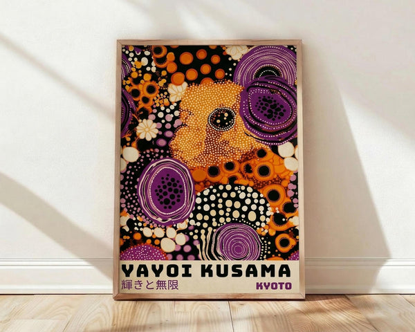 May include: Framed poster with colorful abstract dot pattern artwork leaning against white wall on light wooden floor. Features vibrant orange, purple, black and white circular designs with polka dots in Kusama's signature style. Text at bottom reads "YAYOI KUSAMA" with Japanese characters and "KYOTO". Natural wood frame, soft lighting creates gentle shadows on textured white wall.