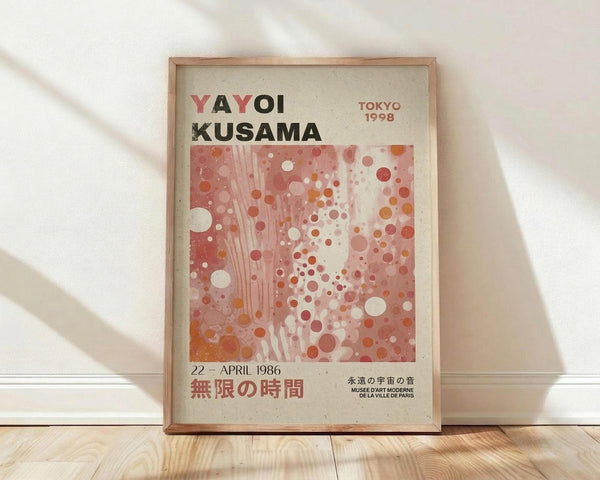May include: Framed exhibition poster with abstract dotted artwork in coral, pink and white tones, featuring Japanese text and "YAYOI KUSAMA" at top, "TOKYO 1998" and museum details, displayed against white textured wall on light wood floor with natural lighting casting soft shadows across the scene.