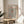 May include: Framed art print with black polka dot pattern on cream background, featuring red circular elements and "YAYOI KUSAMA" text with Japanese characters. Print displayed on round wooden side table next to beige upholstered chair, with plant leaves visible in soft natural lighting.