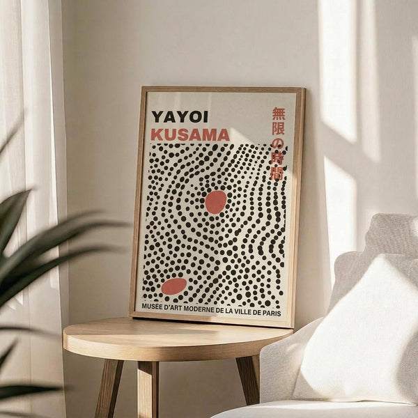 May include: Framed art print with black polka dot pattern on cream background, featuring red circular elements and "YAYOI KUSAMA" text with Japanese characters. Print displayed on round wooden side table next to beige upholstered chair, with plant leaves visible in soft natural lighting.