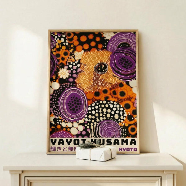 May include: Framed art print leaning against white wall on light wooden console table with small white gift box. Artwork features vibrant abstract design with orange, purple, black and white circular patterns and dots in contemporary style. Japanese text visible at bottom. Natural lighting creates soft shadows on minimalist interior setting.