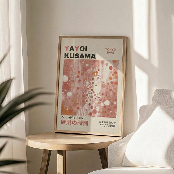 May include: Framed Yayoi Kusama exhibition poster featuring abstract polka dot pattern in coral, pink and orange tones with Japanese text, displayed on round wooden side table next to beige upholstered chair with plant leaves visible in bright, minimalist room setting
