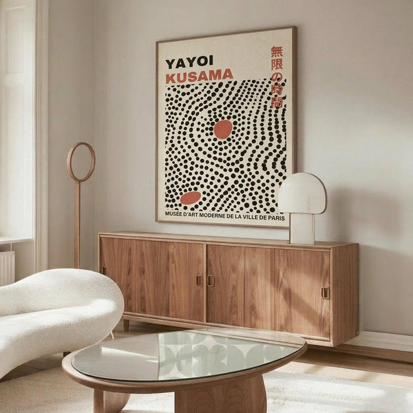 May include: Framed Yayoi Kusama exhibition poster with black polka dot pattern and red accents displayed on wooden sideboard in modern living room. Shows cream-colored furniture, glass coffee table, floor lamp, and neutral wall colors. Poster features spiral dot design with Japanese text and museum information.