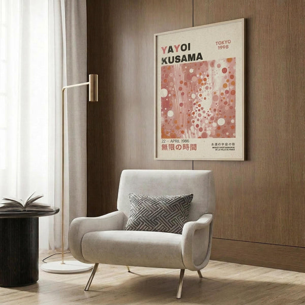 May include: Beige framed poster featuring Yayoi Kusama exhibition text and abstract polka dot pattern in coral, pink and white tones, displayed on wood paneled wall above cream upholstered armchair with geometric patterned pillow, beside brass floor lamp and dark round side table with open book