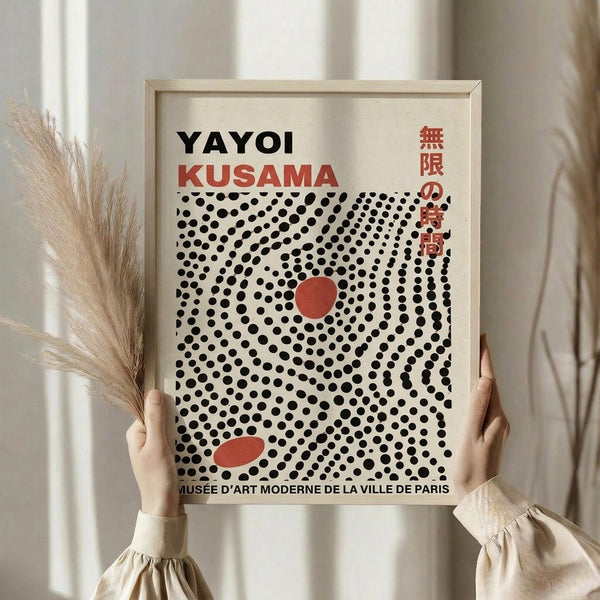 May include: Hands holding a framed art print featuring black polka dots in a spiral pattern with red circular accents on cream background. Text reads "Yayoi Kusama" with Japanese characters and "Musée d'Art Moderne de la Ville de Paris" at bottom. Light wood frame, neutral beige clothing and dried pampas grass visible in soft, minimalist setting with white curtains.