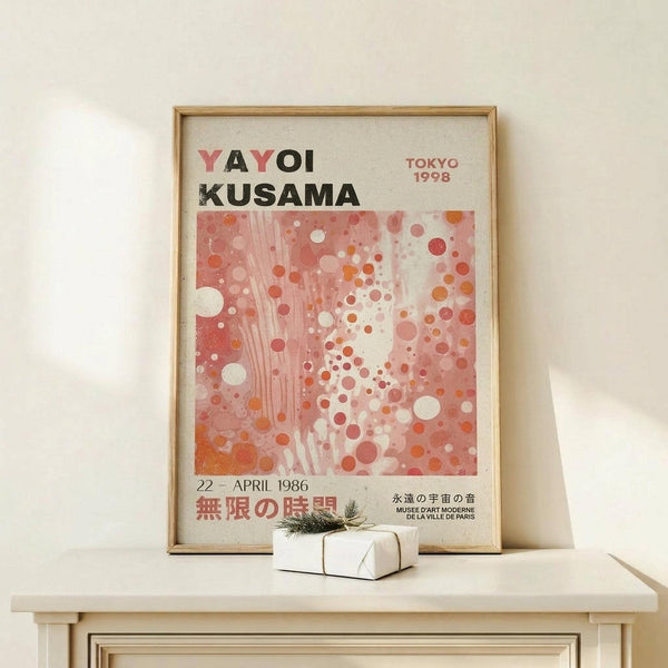 May include: Framed exhibition poster for Yayoi Kusama featuring pink and coral polka dot pattern artwork, displayed on light wood console table with small white gift box, against cream wall. Wood frame complements minimalist styling.
