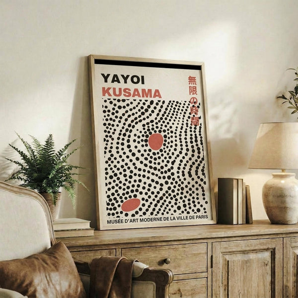 May include: Framed art print displayed on rustic wooden sideboard in neutral living room setting. Poster features black polka dot pattern on cream background with red circular elements and "YAYOI KUSAMA" text at top, referencing Musée d'Art Moderne de la Ville de Paris. Scene includes fern plant, ceramic vase, books, and beige upholstered chair with brown leather cushion.