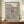 May include: Framed exhibition poster featuring Yayoi Kusama's name in black and red text with Japanese characters, displaying a pattern of black dots arranged in swirling formations with two red oval shapes on cream background. The wooden-framed artwork sits on a walnut credenza beside a white ceramic vase and stacked books in a bright room with white paneled walls.