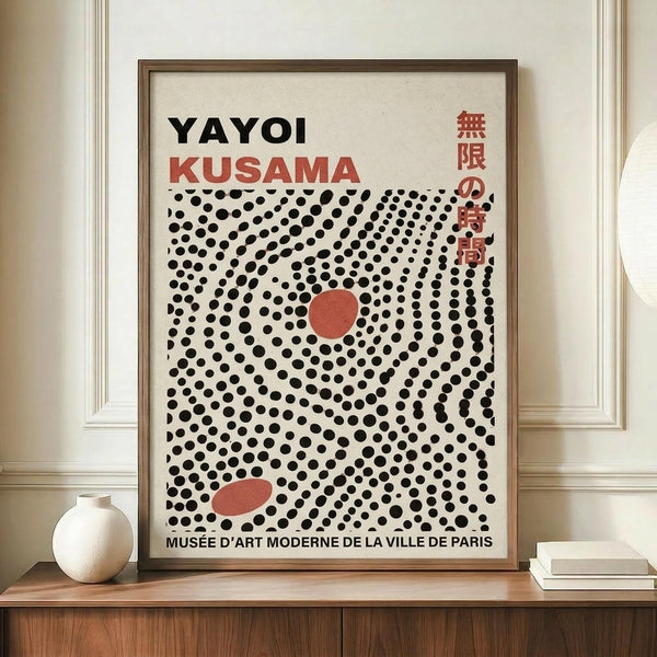 May include: Framed exhibition poster featuring Yayoi Kusama's name in black and red text with Japanese characters, displaying a pattern of black dots arranged in swirling formations with two red oval shapes on cream background. The wooden-framed artwork sits on a walnut credenza beside a white ceramic vase and stacked books in a bright room with white paneled walls.