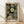 May include: Framed art print leaning against white wall on light wood floor with soft window light shadows. The poster features dotted circular patterns in cream and white on dark green background, resembling stylized flowers or spheres. Text reads "YAYOI KUSAMA" with Japanese characters and "KYOTO" below. Natural wood frame surrounds the vintage-style botanical artwork with pointillism technique.