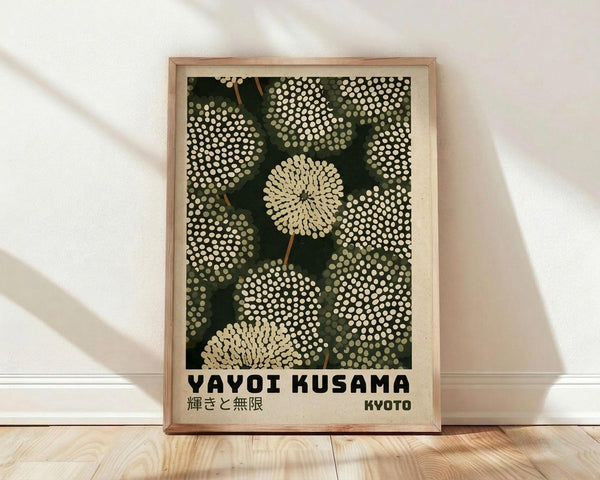 May include: Framed art print leaning against white wall on light wood floor with soft window light shadows. The poster features dotted circular patterns in cream and white on dark green background, resembling stylized flowers or spheres. Text reads "YAYOI KUSAMA" with Japanese characters and "KYOTO" below. Natural wood frame surrounds the vintage-style botanical artwork with pointillism technique.