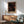 May include: Framed poster with black background featuring orange, cream and rust colored dots in cosmic pattern, displayed above wooden sideboard with white ceramic lamp. Mid-century modern living room setting with sage green upholstered armchair, round wood coffee table, and natural sunlight casting shadows on white wall.