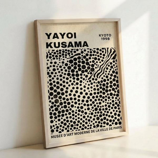 A framed poster print featuring black polka dots of varying sizes on cream background with wavy lines cutting through the pattern. The top displays "YAYOI KUSAMA" and "KYOTO 1998" in bold black text, with "MUSEE D'ART MODERNE DE LA VILLE DE PARIS" at bottom. The artwork is displayed in a light wood frame leaning against a white wall on a white surface, creating subtle shadows.