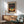 May include: Framed poster featuring a stylized tree with colorful dotted foliage against a gradient sky, displayed above a wooden sideboard with white ceramic lamp in a modern living room setting with mint green upholstered chair and round coffee table, warm natural lighting creating shadows on white walls