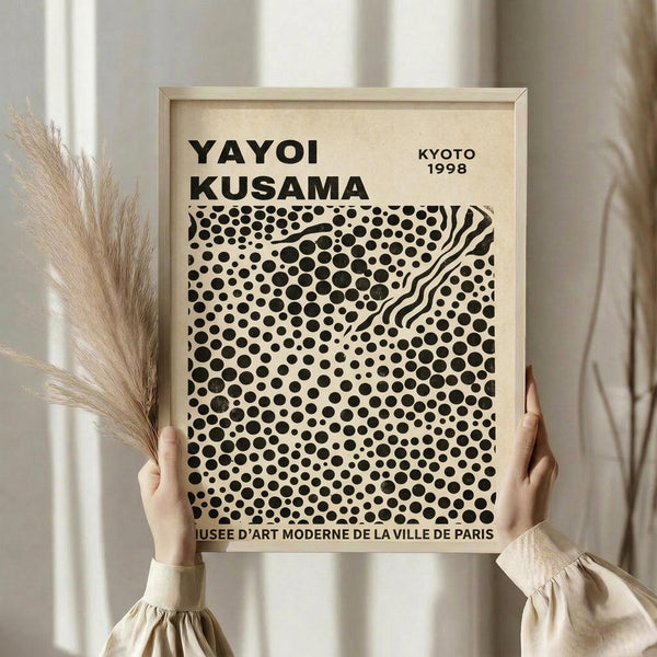 May include: Person in cream ruffled sleeves holding framed museum poster with black polka dot pattern on cream background. Light wood frame against neutral wall with dried pampas grass visible to left. Exhibition poster shows dense circular dots with flowing curved lines, museum text at bottom.