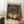 May include: Framed poster featuring a silhouetted tree with colorful polka dot foliage against a vibrant sunset sky, displaying "YAYOI KUSAMA" at top and "TOKYO 1998" at bottom with Japanese characters. The artwork shows Kusama's signature dot style in blues, oranges, purples, and yellows. Frame leans against white paneled wall next to wooden side table with coffee cup, positioned on white textured rug beside beige upholstered furniture.