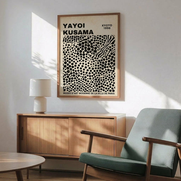 May include: Framed vintage museum poster featuring black polka dot pattern on cream background, displayed above wooden sideboard with white ceramic lamp in modern living room setting with mint green upholstered armchair and natural sunlight streaming across white walls.