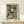 May include: Framed poster print featuring green background with white dotted circular chrysanthemum-like flowers and Japanese text reading "YAYOI KUSAMA" and "KYOTO" at bottom. Natural wood frame sits on light wooden console table next to small wrapped white gift box with pine sprig. Clean white wall background creates minimalist styling.