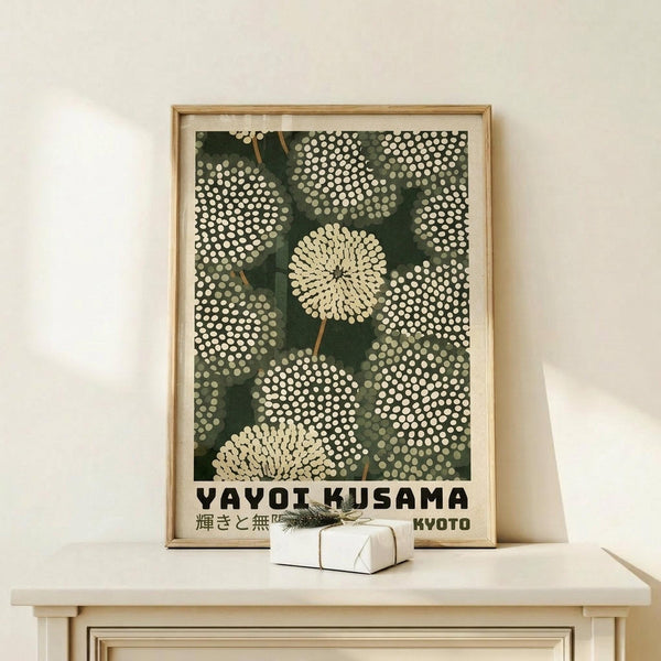 May include: Framed poster print featuring green background with white dotted circular chrysanthemum-like flowers and Japanese text reading "YAYOI KUSAMA" and "KYOTO" at bottom. Natural wood frame sits on light wooden console table next to small wrapped white gift box with pine sprig. Clean white wall background creates minimalist styling.