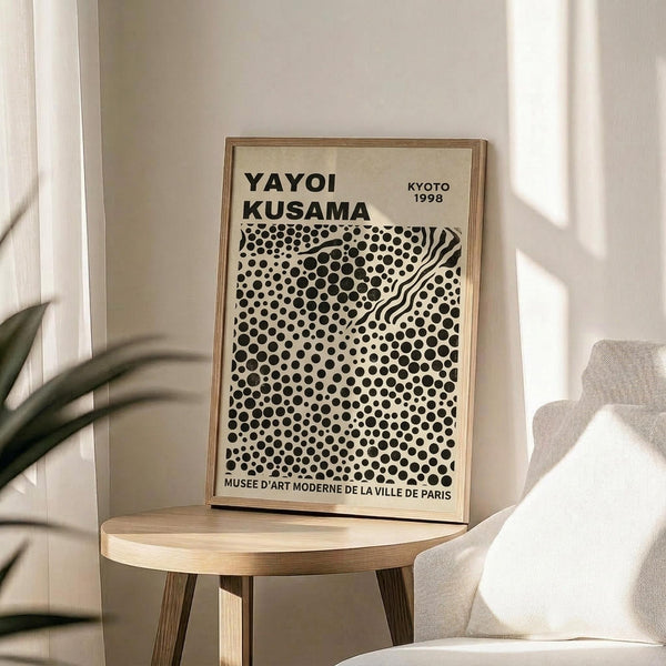 May include: Framed poster with black polka dot pattern and wavy lines on cream background, displayed on light wood round side table next to beige upholstered chair. Natural lighting creates soft shadows on white wall behind. Green plant leaves visible in corner. The artwork features dense circular dots in varying sizes with flowing curved lines creating visual movement across the composition.