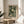 May include: Framed art print featuring Yayoi Kusama's polka dot artwork with white dots on deep green background and Japanese text, displayed on round wooden side table next to beige upholstered chair. Natural light streams across white wall creating soft shadows, with plant leaves visible in corner of minimalist room setting.