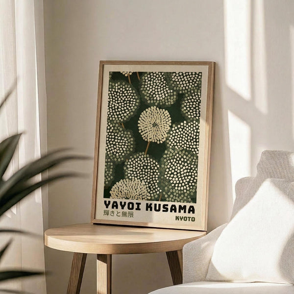 May include: Framed art print featuring Yayoi Kusama's polka dot artwork with white dots on deep green background and Japanese text, displayed on round wooden side table next to beige upholstered chair. Natural light streams across white wall creating soft shadows, with plant leaves visible in corner of minimalist room setting.