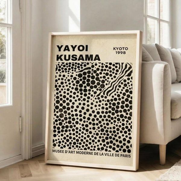 May include: Framed art print leaning against white wall next to beige upholstered chair in bright living room. Poster features black polka dots of varying sizes on cream background with wavy striped pattern cutting diagonally through center. Bold black text at top reads artist name, location and year. Museum attribution at bottom. Light wood frame, natural lighting from large windows.