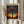 May include: Hands holding a framed art print featuring a stylized tree with colorful polka dot foliage against a vibrant sunset sky. The artwork shows Kusama's signature dotted style with the tree's canopy made of multicolored circles in blues, oranges, purples and greens. Text reads "YAYOI KUSAMA" at top and "TOKYO 1998" with Japanese characters at bottom. Light wood frame, neutral background with dried wheat stems visible on left side.