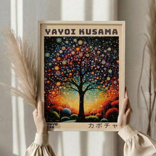 May include: Hands holding a framed art print featuring a stylized tree with colorful polka dot foliage against a vibrant sunset sky. The artwork shows Kusama's signature dotted style with the tree's canopy made of multicolored circles in blues, oranges, purples and greens. Text reads "YAYOI KUSAMA" at top and "TOKYO 1998" with Japanese characters at bottom. Light wood frame, neutral background with dried wheat stems visible on left side.