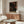 May include: A stylized room interior with wooden sideboard and white sculptural chair. Above the furniture hangs a framed poster featuring a black background with scattered circles and dots in warm orange, red, and cream tones, creating a cosmic or cellular pattern. The poster has Japanese text beneath the artwork. A modern table lamp and glass coffee table complete the minimalist, contemporary setting with white walls.