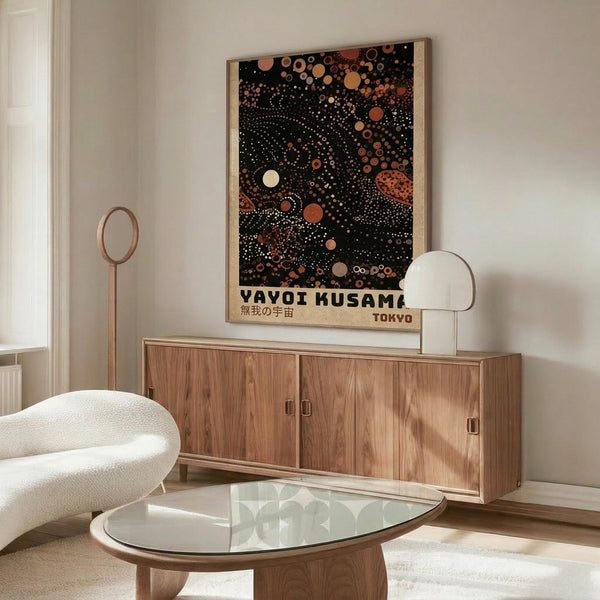 May include: A stylized room interior with wooden sideboard and white sculptural chair. Above the furniture hangs a framed poster featuring a black background with scattered circles and dots in warm orange, red, and cream tones, creating a cosmic or cellular pattern. The poster has Japanese text beneath the artwork. A modern table lamp and glass coffee table complete the minimalist, contemporary setting with white walls.