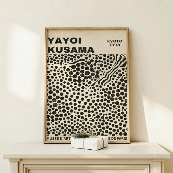 May include: Framed museum exhibition poster featuring Yayoi Kusama's signature black polka dot pattern on cream background, displayed on light wood console table against white wall. Small wrapped gift box with greenery sits beside the thin wooden frame.