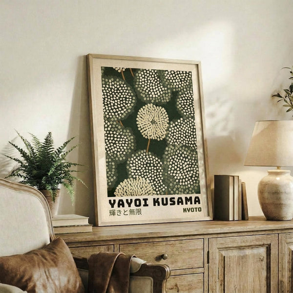 May include: Framed poster with green background and white polka dot pattern resembling dandelion seed heads, displayed on rustic wooden sideboard alongside fern plant, books, and ceramic vase in neutral-toned living room setting with cream armchair visible.