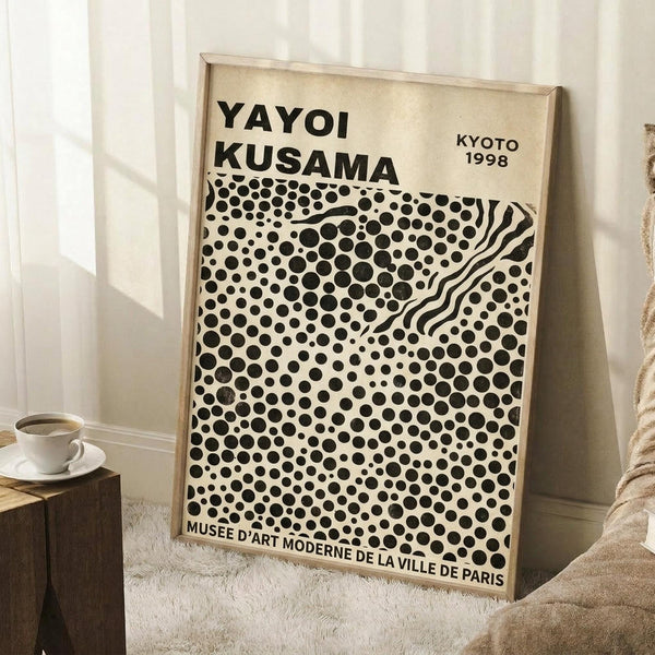 May include: A framed exhibition poster featuring black polka dots of varying sizes on cream background with wavy lines creating movement through the pattern. The poster displays "YAYOI KUSAMA" and "KYOTO 1998" at the top, with "MUSEE D'ART MODERNE DE LA VILLE DE PARIS" at bottom. Light wood frame leans against white wall on textured cream rug, with coffee cup on wooden block nearby and soft lighting creating shadows.