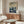 May include: Framed poster displaying an abstract landscape with flowing waves in blue and cream tones above a field of white mushrooms and dark plants against a starry sky. Shown in a modern living room with light wood sideboard, white curved chair, glass coffee table, and minimalist copper floor lamp on polished floors.