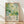May include: Framed art print leaning against white wall on light wood floor with natural sunlight casting shadows. Abstract botanical artwork features overlapping green circles, dots, and leaf shapes in various shades of sage, teal, and yellow-green. Text reads "YAYOI KUSAMA" with Japanese characters and "JAPAN 1987" below the dreamy, organic composition in thin wood frame.