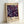 May include: Framed museum exhibition poster featuring Yayoi Kusama's colorful floral artwork on dark purple background with scattered daisy-like flowers in blues, oranges, and pinks. Text shows dates "28. APRIL - 14. MAI 1984" and "MUSÉE D'ART MODERNE DE LA VILLE DE PARIS" on cream colored background. Light wood frame leans against white wall.