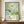 May include: Framed art print with abstract green and teal botanical design featuring organic shapes, circles, and flowing forms in watercolor style. White text reads "YAYOI KUSAMA" with "JAPAN 1987" and Japanese characters below. Print displayed on wooden sideboard next to white ceramic vase and stacked books in room with white paneled walls.