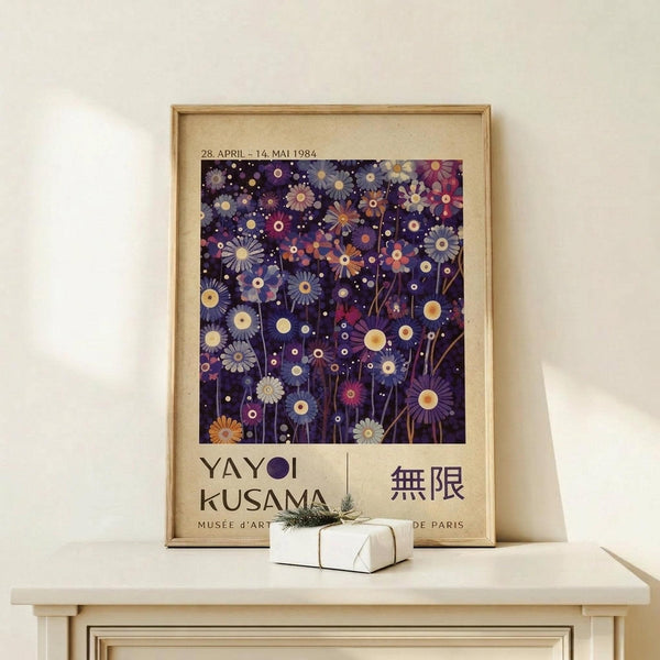 May include: Framed vintage exhibition poster with dark purple background featuring colorful abstract flowers and circles in blues, pinks, and yellows. Text reads "YAYOI KUSAMA" and shows museum details. Natural wood frame sits on light wooden console table next to small wrapped white gift box with pine sprig against cream wall.
