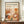 May include: Framed art print displayed on wooden sideboard featuring abstract botanical illustration in warm orange, brown, and cream tones with stylized plants, circles, and dotted patterns. Text reads "YAYOI KUSAMA" with Japanese characters and "JAPAN 1987." Small white ceramic vase and stacked books sit nearby on the furniture surface against white wainscoted wall.