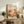 May include: Framed poster with cream background featuring "YAYOI KUSAMA" text, Japanese characters, and "JAPAN 1987" in orange lettering. Abstract botanical illustration shows warm-toned organic shapes, polka-dotted mushrooms, and stylized plants in oranges, browns, and greens. Natural wood frame sits on rustic wooden sideboard alongside fern plant, ceramic vase, and stacked books in neutral-toned living room setting.