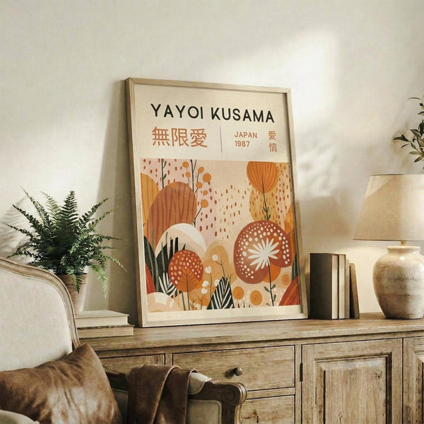 May include: Framed poster with cream background featuring "YAYOI KUSAMA" text, Japanese characters, and "JAPAN 1987" in orange lettering. Abstract botanical illustration shows warm-toned organic shapes, polka-dotted mushrooms, and stylized plants in oranges, browns, and greens. Natural wood frame sits on rustic wooden sideboard alongside fern plant, ceramic vase, and stacked books in neutral-toned living room setting.
